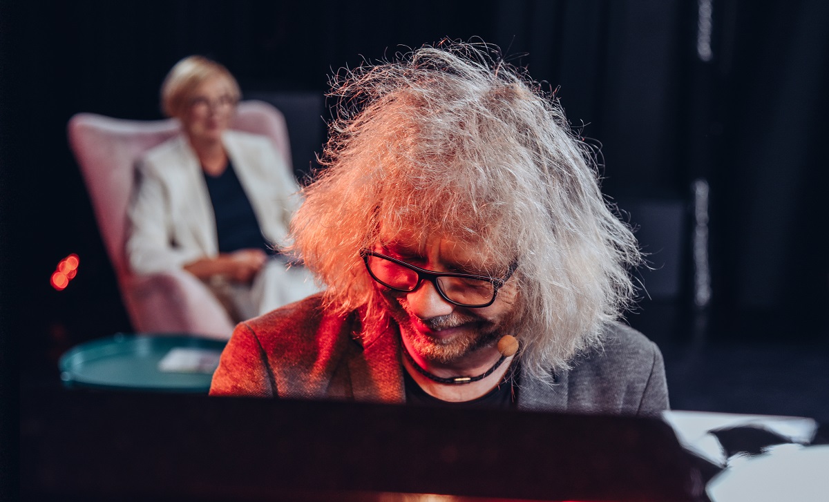 Na zdjęciu Joachim Mencel z siwą rozczorchaną fryzurą w okularach. Uśmiecha się grając na fortepianie. Za nim w tle kobieta w białym kostiumie siedzi na różowym fotelu.