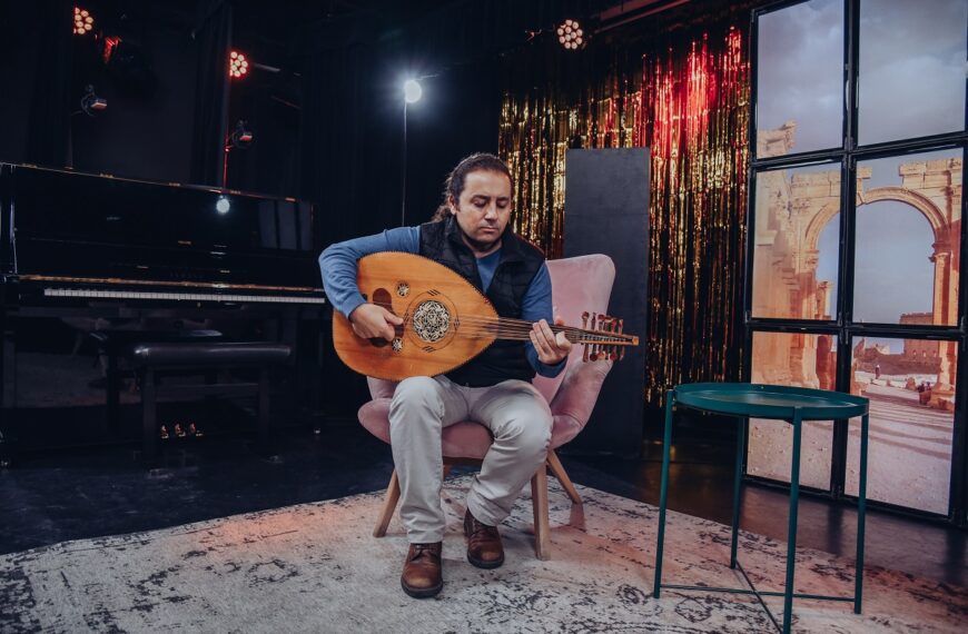 Na zdjęciu Wassim Ibrahim siedzi na fotelu i gra na lutni arabskiej zwanej Ud. Po prawej mały okrągły stolik. Za nim plan telewizyjny