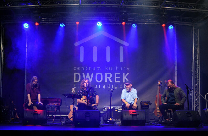 Na scenie czworo muzyków z zespołu Hard Times gra koncert. Oświetleni niebieskim światłem. W tle logotyp instytucji.