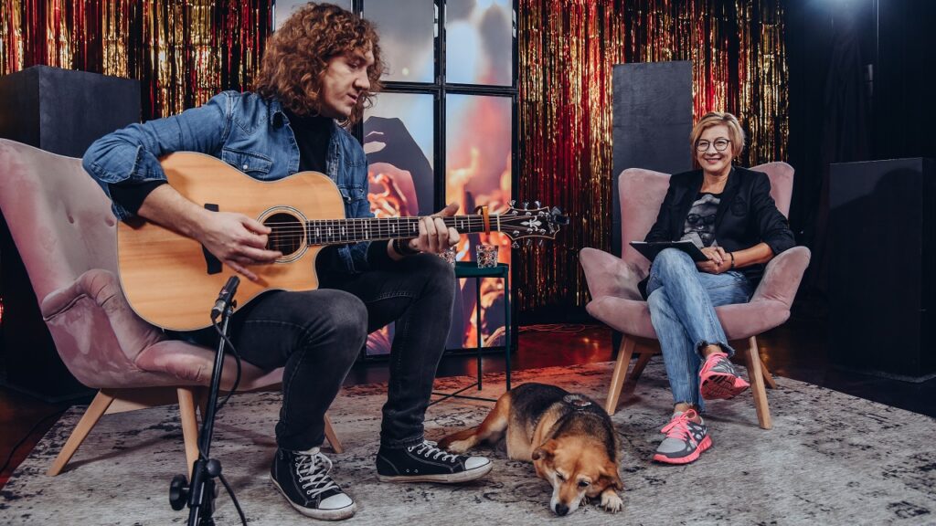 Od lewej Łukasz Wiśniewski gra na gitarze, na podłodze leży pies. Po prawej na fotelu siedzi kobieta.