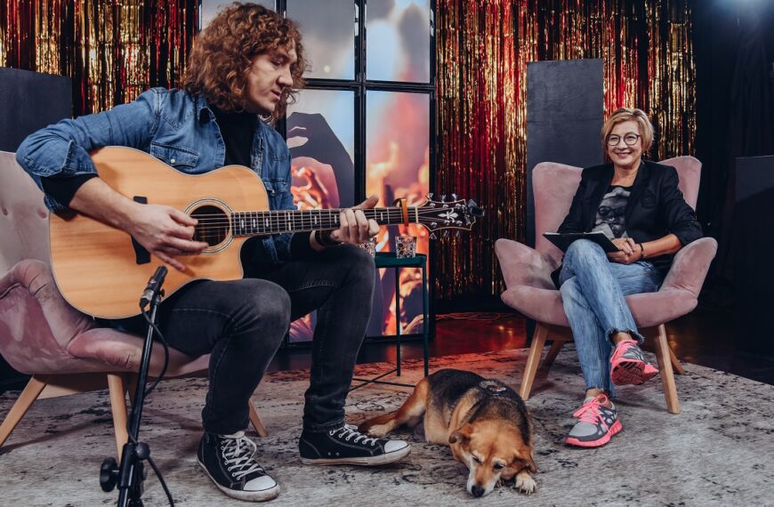 Od lewej Łukasz Wiśniewski gra na gitarze, na podłodze leży pies. Po prawej na fotelu siedzi kobieta.