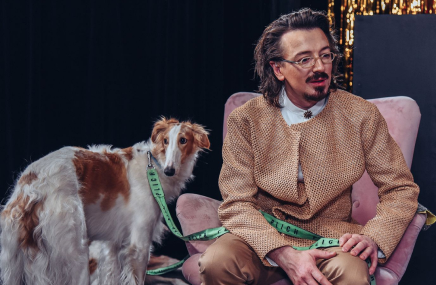 Po prawej mężczyzna (PLICH) w beżowym garniturze siedzi na fotelu. Po prawej stoi pies - chart.