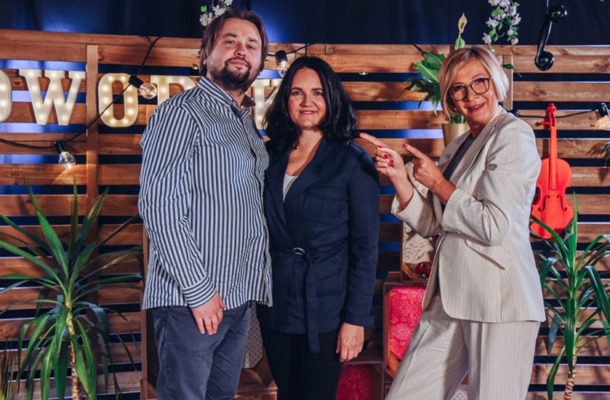 Na planie zdjęciowym po lewej goście: kobieta i mężczyzna z zespołu Cracow Singers. Po prawej prowadząca wskazuje na nich rękami.