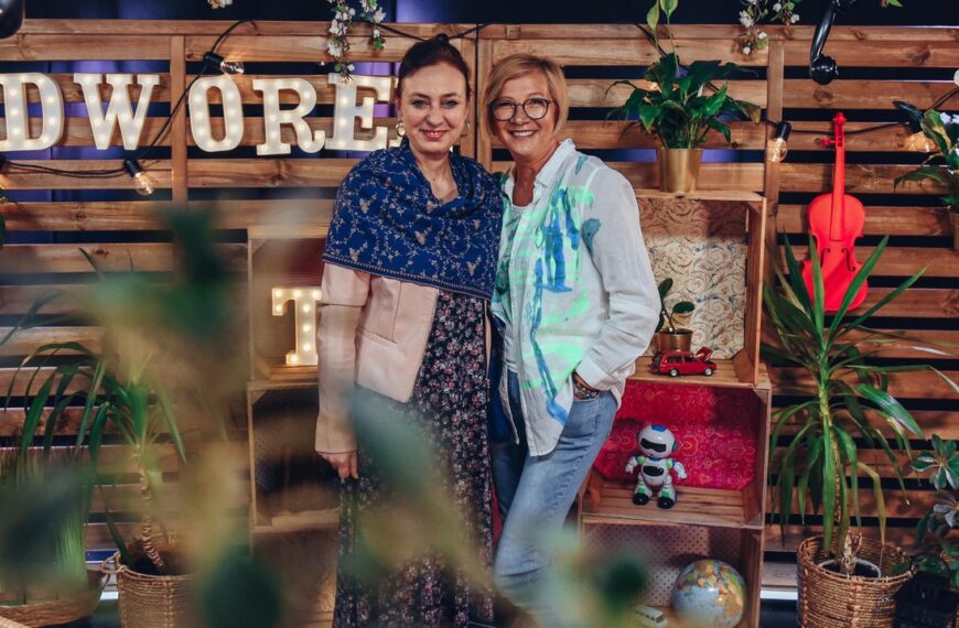 Dwie uśmiechnięte kobiety pozują do zdjęcia. Romana Agnel po lewej. W tle napis DWOREK TV