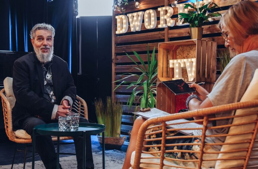 Po lewej stronie siedzi uśmiechnięty Jorgos Skolias z brodą. Po prawej odwrócona tyłem kobieta siedząca na krześle.