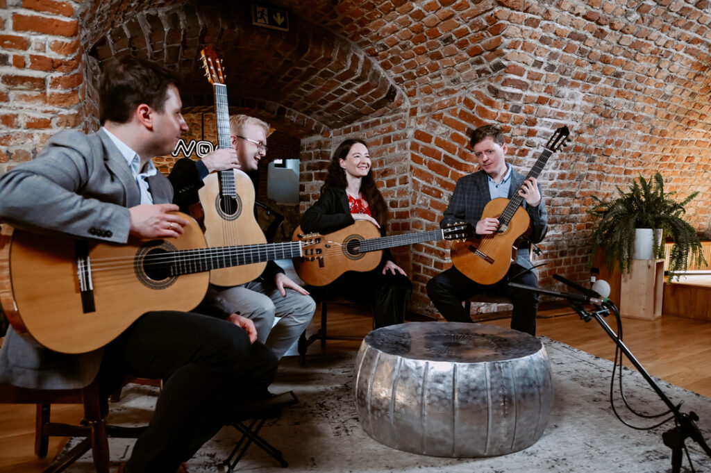 Na zdjęciu czterech muzyków z zespołu Cracow Guitar Quartet podczas wywiadu w Dworek TV. Na zdjęciu widać trzech mężczyzn i kobietę grających na gitarach.