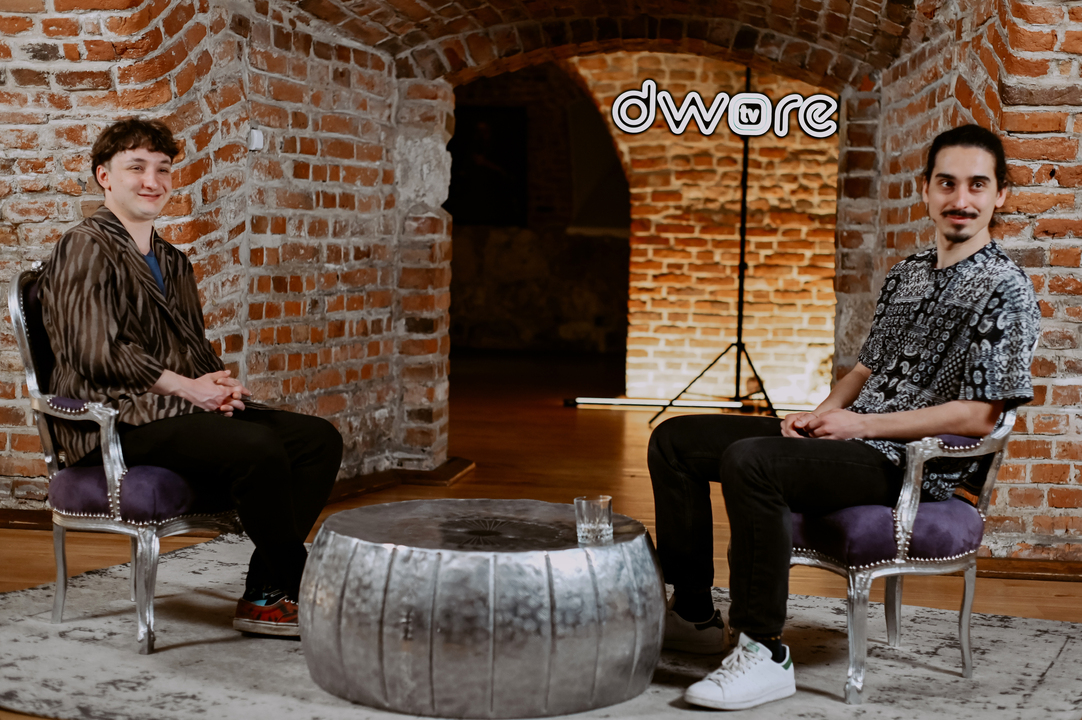 Wojciech Szumański i Jakub Byczek podczas nagrania w Dworek TV. Mężczyźni siedzą w piwnicy Dworku, z tyłu widzimy neon.
